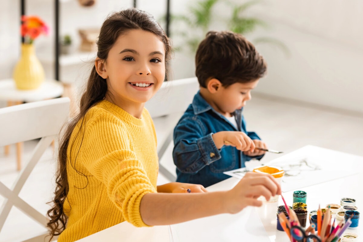 Cheerful sister looking at camera while sitting at table and drawing together with brother