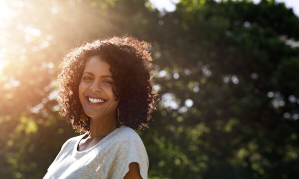 Portrait of a young woman with curly hair smiling while standing outside in a park on a sunny summer afternoon