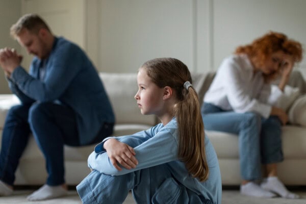 Unhappy girl suffering from loneliness while her parents sitting on background by sides on sofa after quarrel, selective focus on child